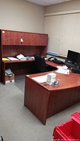 Wide angle of office desk with curved front and overhead cabinet. Desk is cherry laminate particle board with scratches. Overhead cabinet mounted above desk with two doors.