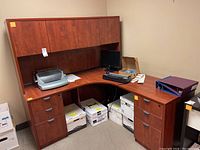 Brown wooden corner desk setup with office items including a keyboard, monitor, and paper binding machine on top, and multiple stacks of paper below the desk. The desk has six drawers and an overhead storage unit with cabinets.