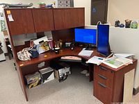 L-shaped brown particleboard desk with overhead cabinets, desktop clutter including dual monitors, keyboard, and office supplies, showing overall desk structure and condition.