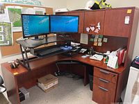 Full view of brown particleboard corner desk with dual monitors on a black stand, keyboard tray, and overhead storage cabinet.