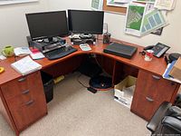 L-shaped brown particleboard wooden office desk with dual monitors and office accessories on top, showing desk shape and surface condition