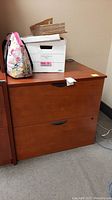 Full front view of the wood veneer covered particleboard 2-drawer file cabinet with box and bag on top, shows medium brown finish with black drawer handles, scratches are visible