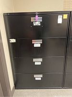 Full frontal view of the closed black lateral filing cabinet showing four drawers, key lock, and silver horizontal handles. Some scratches visible on surface.
