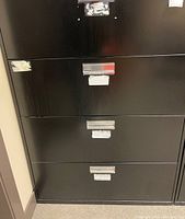 Close-up front view of black lateral filing cabinet with four drawers, showing scratches and wear on surfaces and drawer handles.