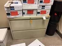Cream lateral filing cabinet with two horizontal drawers, placed on floor next to a black standing dispenser and several boxes on top. Cabinet has a lock with orange key tag.