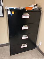 Front view of black 4-drawer filing cabinet with labels on drawers and some items on top