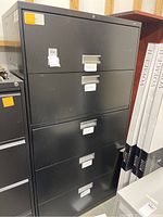 Black metal 5-drawer filing cabinet with label holders and keys, showing scratches and wear on surface.