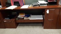 Front view of wooden office desk with papers and folders on top and shelf; metal handles on storage compartments visible