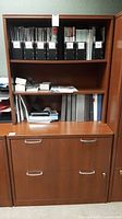 Front view of the particleboard office cabinet showing two shelves with visible office binders (not included), two drawers with metal handles, and wood-grain finish with scratches.