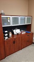 Full front view of office cabinet with various office supplies on top, showing glass doors and wood finish.