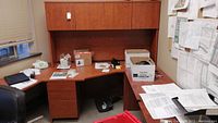 Full view of L-shaped laminate office desk with drawers and overhead storage cabinets in an office setting with papers and boxes on the surface.