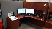 L-shaped office desk in cubicle with dual monitors, phone, paperwork, showing overall view of desk and layout.