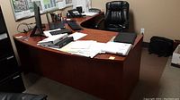 Full view of L-shaped office desk in an office with visible papers, monitors and chair showing scratches on desk surface.