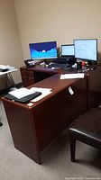 L-shaped office desk in room with office items on top including computer monitors, keyboard, mouse, papers, and chair beside desk.
