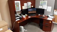 Photo of an L-shape corner office desk covered with a reddish-brown wood grain particleboard surface. Desk has a monitor, keyboard, printer, and office supplies (not included in lot). Hutch above desk for added storage. Desk positioned against corner wall with plans pinned on hutch.