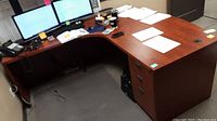 View of the L-shaped desk with office items placed on top, showing the woodgrain finish, surface wear, and cable grommet hole.
