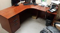L-shaped particleboard office desk with a cherry wood laminate surface, showing the left side with three drawers and corner workspace with visible scratches.