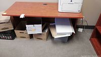 View of the full desk showing the laminate surface, scratches, and cable management hole near back right corner. The table is set against a wall and sitting on carpet floor with boxes underneath.