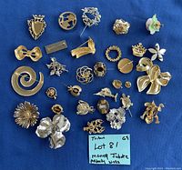 A collection of 27 vintage jewelry pins arranged on a blue background showing various styles and colors, including metallic gold and silver tones.