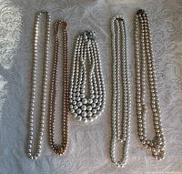 Lot of five pearl necklaces arranged on white fabric showing variation in pearl size, color, and clasp style.