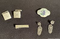 Photo showing a pair of square cufflinks with starburst design, a rectangular tie clip, an enamel floral brooch, and a pair of dangle earrings with floral motifs and chains.