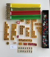 Top-down image showing laid out Mah-Jongg tiles separated by collection type, scoring sticks in yellow, green and red with brass ends, and a box of multi-colored circular chips.