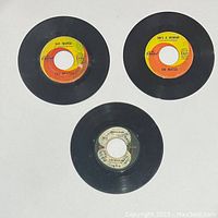 Three 45 RPM Beatles vinyl records with Capitol and Apple Records labels visible, arranged on a white background. Close up of labels shows song titles and label logos.