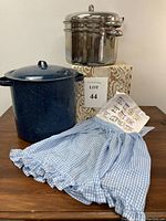 Lot with stainless steel pasta pot stacked on a box, beside a large blue speckled pot and a blue gingham apron displayed in front.