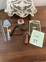 Seven vintage kitchen tools including biscuit cutters, bottle opener, cheese cutter, poultry lancers, heart-shaped mold and springs arranged on wooden surface.