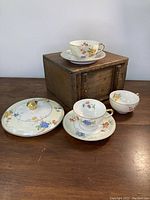 Photo of the entire set with 3 tea cups, 2 saucers and serving bowl lid arranged on a wooden table with a wooden box in the background.