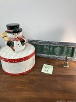 Overall view of the four holiday-themed items on a wooden surface including the snowman cookie jar, wicker hat box, green chalkboard frame, and silver Santa bell.