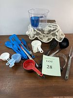 Photo showing all items in lot including measuring cups, kitchen utensils, and tools arranged on a wooden table with lace cloth and a lot number card