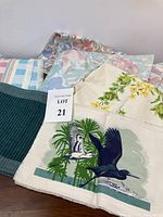 Overview of assorted table linens showing different patterns including multicolored stripes, pastel florals, solid green, and beige linen with bird and ship screen print.