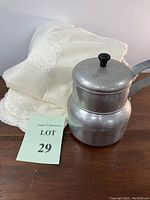 Full view of vintage aluminum double boiler pot and folded off-white lace-edged tablecloth behind it on wooden surface.