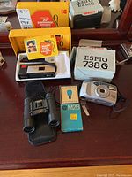 Kodak Pocket Instamatic 20 camera in original yellow box with manual, and Pentax Espio 738G in white box, plus GE flash bulbs and Bushmaster binoculars on wooden surface.