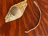 Broken pieces of a 10K gold ring, showing decorative oval face with initials and bent band fragment on wooden surface.