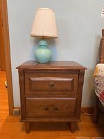 Front view of wood nightstand table with lamp on top, showing drawer pulls and overall style.
