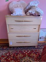 Front view of a light wood dresser with three drawers and gold-colored handles with fabric cushions and textiles on top.