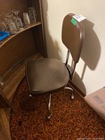 Side angle of the brown retro office chair next to wooden shelving and orange-brown carpet showing vinyl seat and chrome legs with casters.