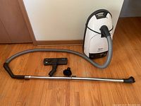Full view of vacuum cleaner, hose, metal extension tube, floor head, and round brush on hardwood floor.