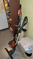 Photo showing two electric weed wackers standing near a shelf in basement-like environment, one Orange Black and Decker, one yellow black Yardworks