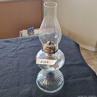 Side view of the clear glass oil lamp showing ribbed base, rounded fuel reservoir, metal burner with wick, and tall glass chimney on a black fabric surface.