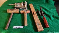 Seven antique hand tools including a metal vice, two wooden mallets, a wooden level with bubble vial, and a manual hand drill with red wooden handle and two hand blades.