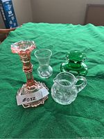 Four assorted glass and crystal pieces arranged on green cloth: pink glass candle holder, small clear vase, green glass vase, clear etched creamer