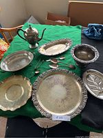 Photo showing entire assortment of silver-plated trays, teapot, and serving utensils arranged on a green cloth.