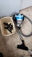 Bissell vacuum cleaner with hose and floor cleaning attachment, basket holding several extra brush accessories on a tiled floor.