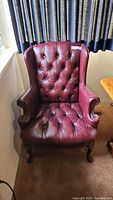 Front view of burgundy leatherette wingback chair showing tufted back and seat, nailhead trim on arms, carved wooden legs.