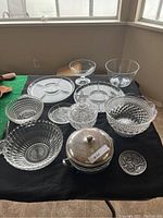 Photo showing 11 items including multiple crystal and glass bowls and plates arranged on a black cloth. Silver topped bowl with lid is centered in front.