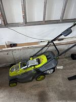 SunJoe electric lawn mower with battery charger placed on garage floor against wall, showing overall mower in green and gray colors with push handle and wheels.