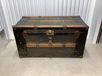 Front view of antique steamer trunk showing black outer material, wooden slats, and central lock mechanism.